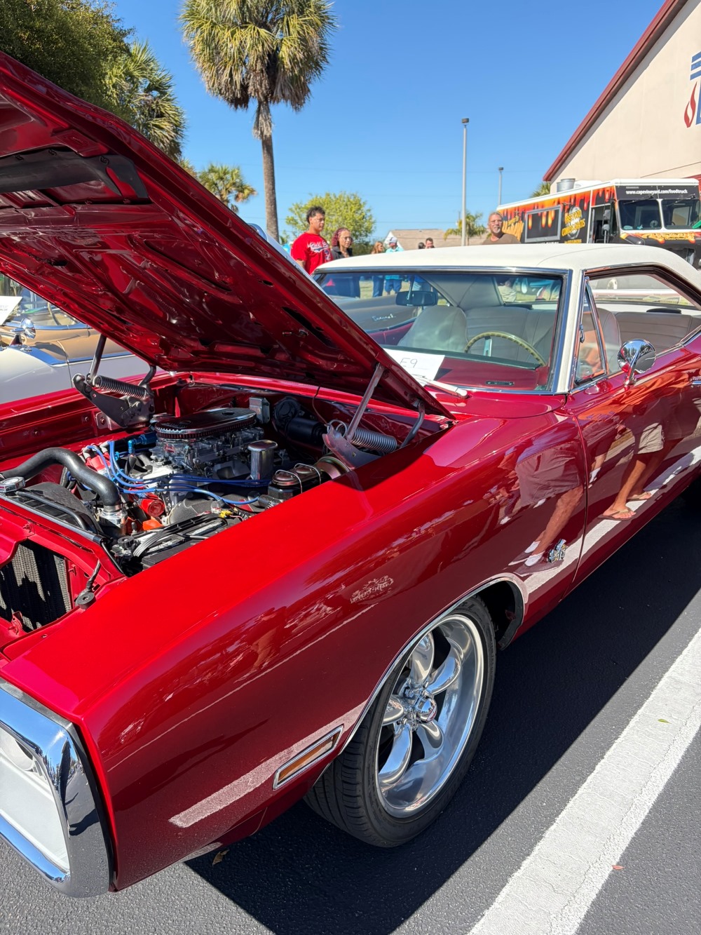 Bright red classic muscle car with its hood open, revealing the engine, parked near palm trees at a car show